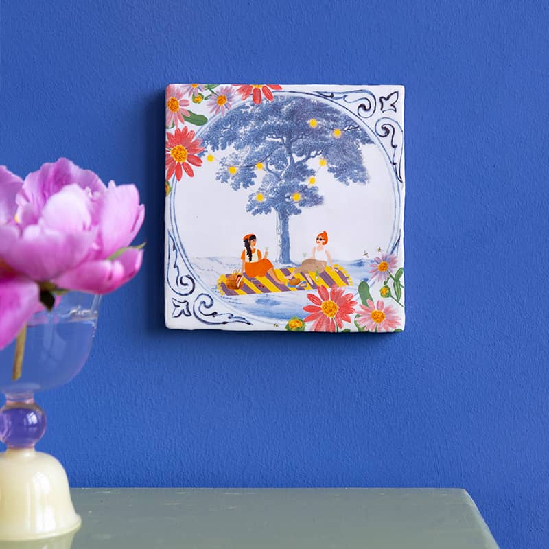 Delfts blauw tegeltje met mensen onder boom, omringd door bloemen