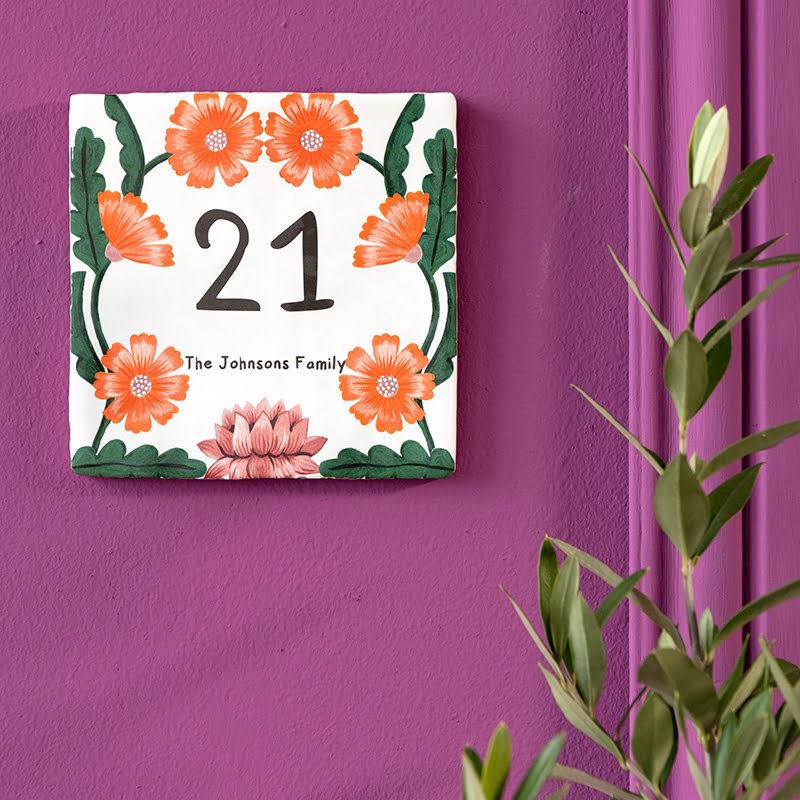 Huisnummer tegeltje met oranje bloemen op paarse muur bevestigd