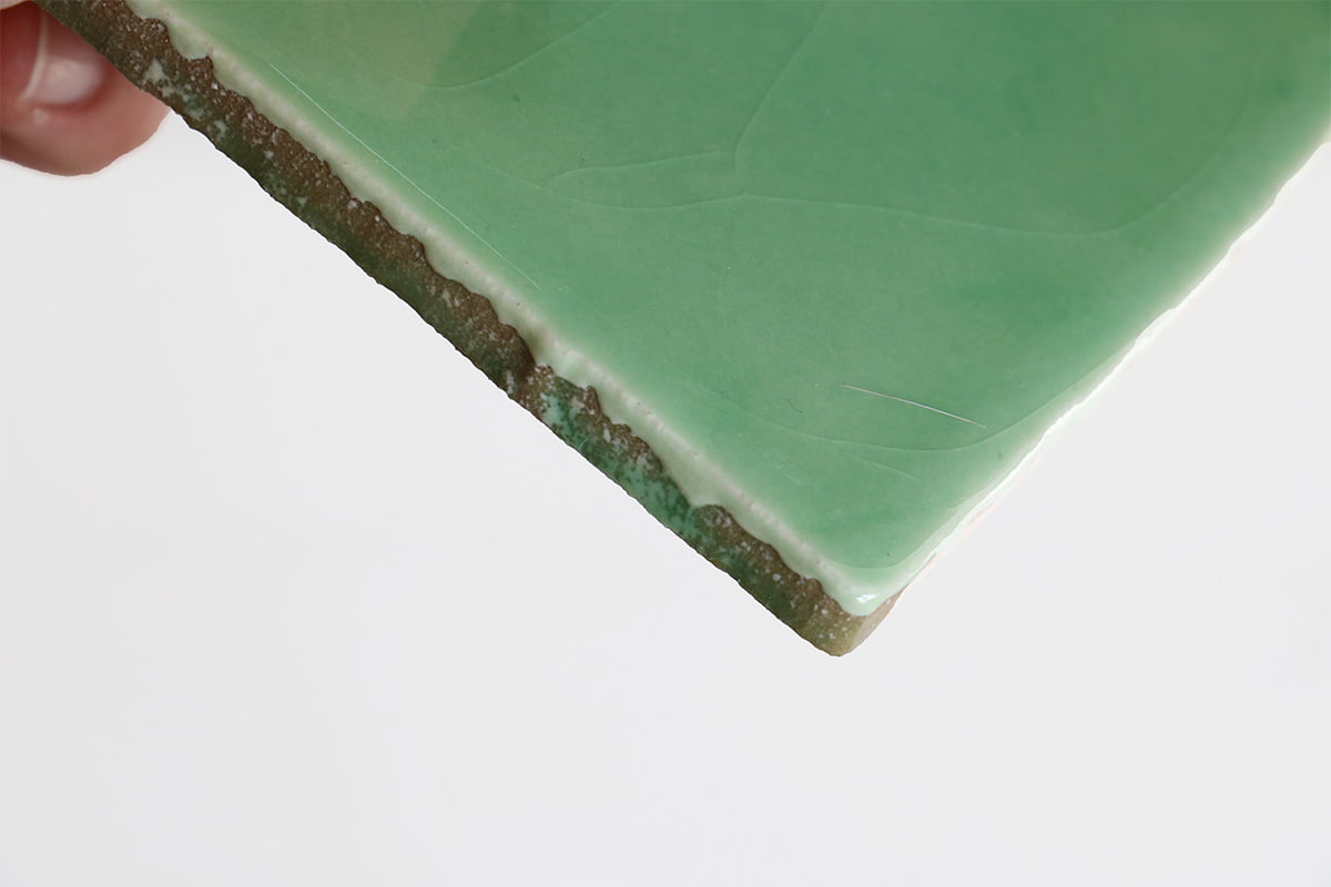 Groen geglazuurd keramisch tegeltje, vastgehouden met vingers, rand zichtbaar