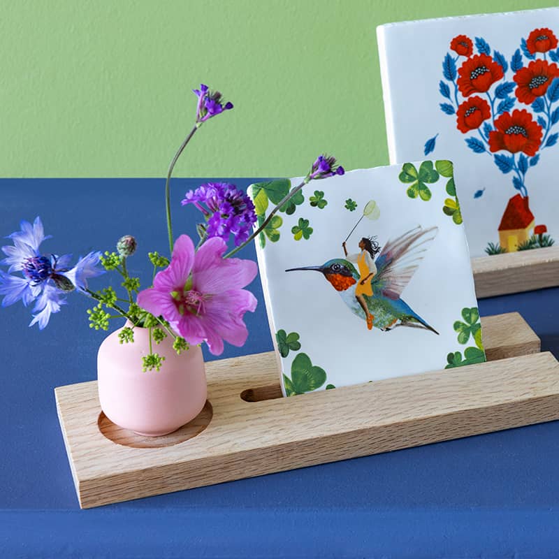 Tegeltje met vogel en persoon, geplaatst tussen bloemen op standaard