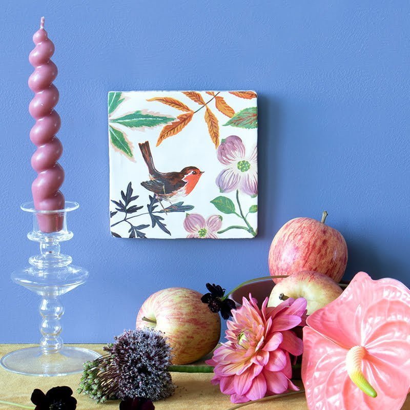 Tegeltje roodborst tussen bloemen, naast roze kaars en fruit