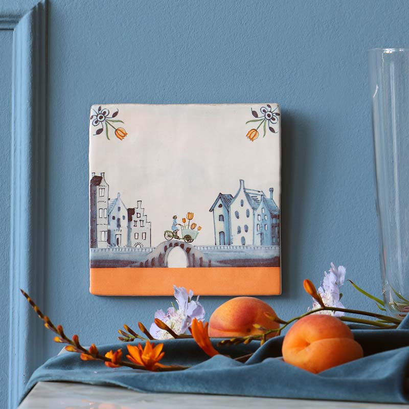 Gepersonaliseerd tegeltje met grachtenhuizen en bloemen, tegen blauwe muur