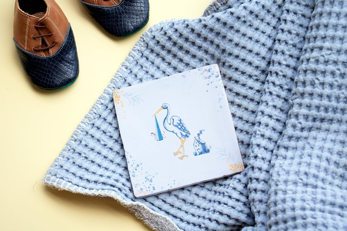 Geboortetegel ooievaar op blauwe deken met babyschoentjes