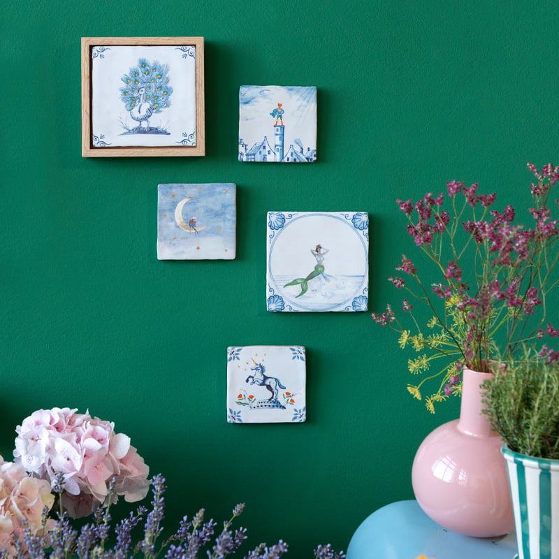 Delfts blauwe tegels verschillende illustraties aan groene muur met bloemen