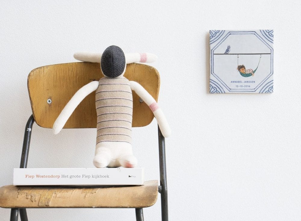 Tegeltje met kindje in hangmat aan muur naast knuffel en boek op stoel