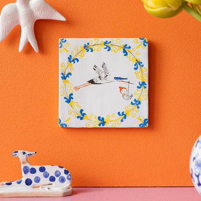 Delfts blauwe geboortetegel met Fiep Westendorop tekening Hallo kleintje met ooievaar die kindje brengt, opgehangen aan een oranje wand