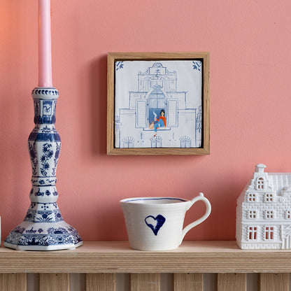 Tegeltje met de gevel van een grachtenpand en meisje dat in het raam koffie drinkt, ingelijst in houten frame