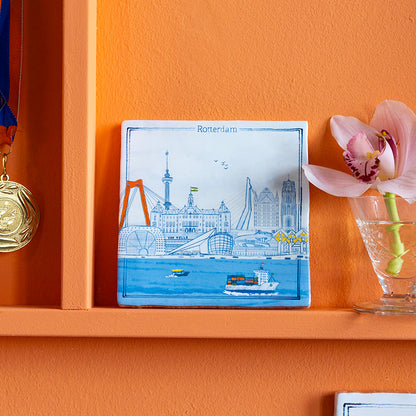 Tegeltje met Delfts blauwe tekening van Rotterdam tegen een oranje wand
